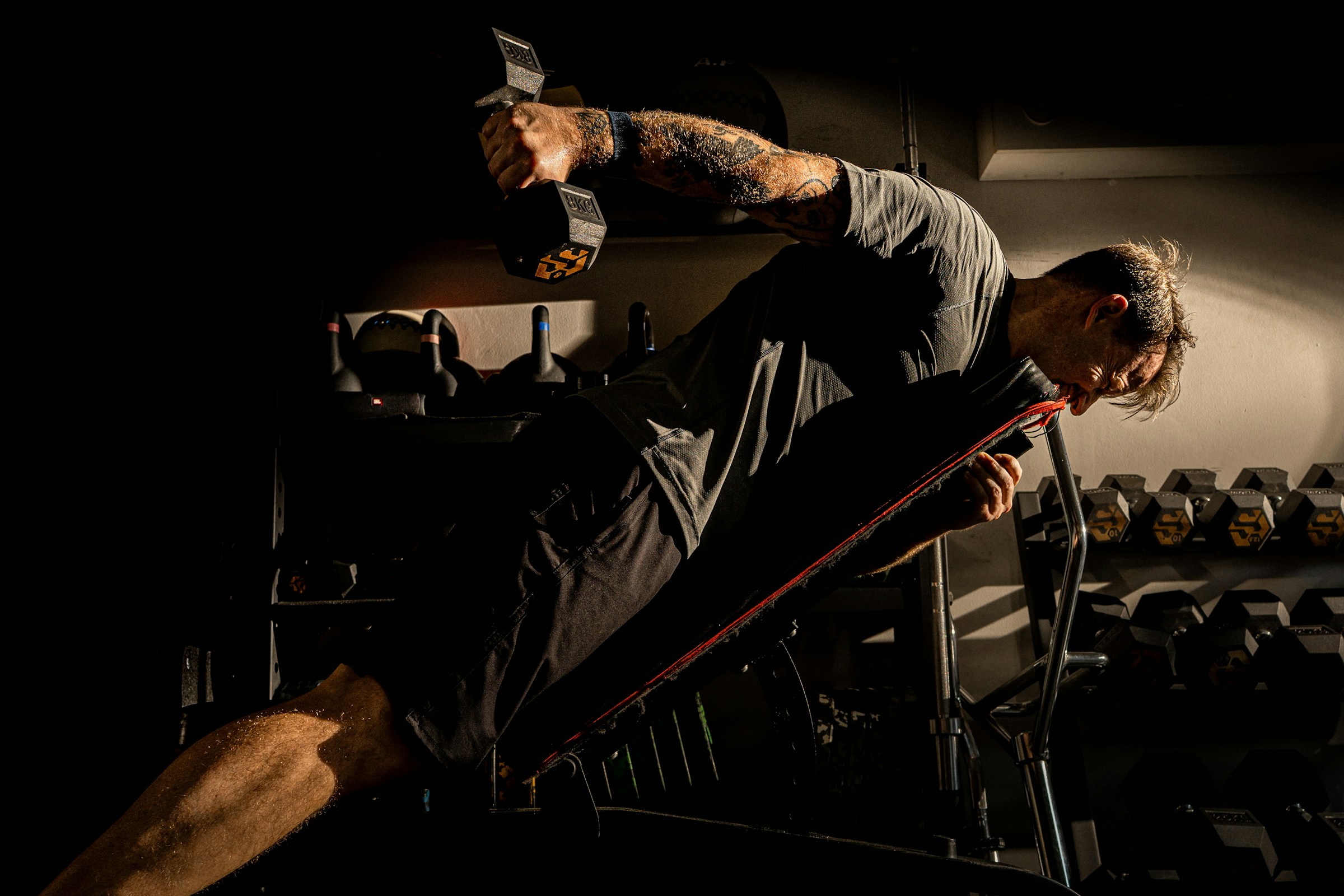 Man mid dumbbell row in a dark home gym, golden rim light